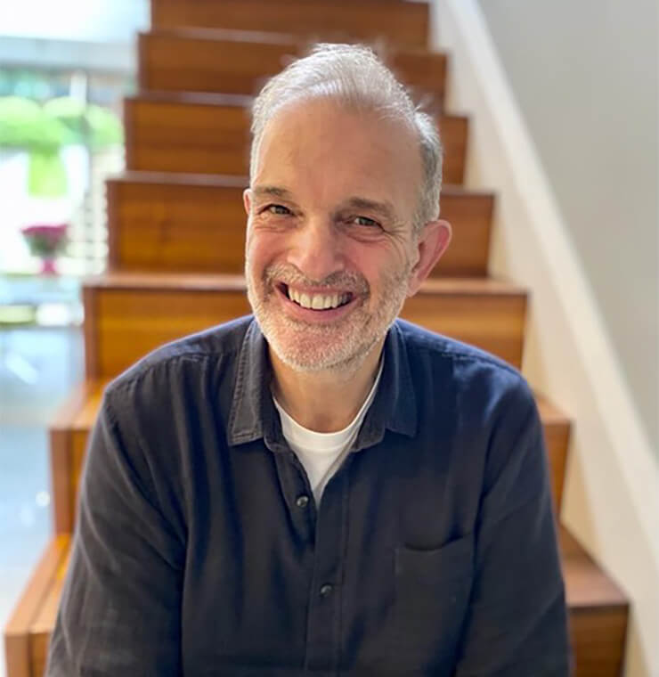 Smiling middle-aged man with short gray hair and beard, wearing a dark shirt, sitting in front of wooden stairs.