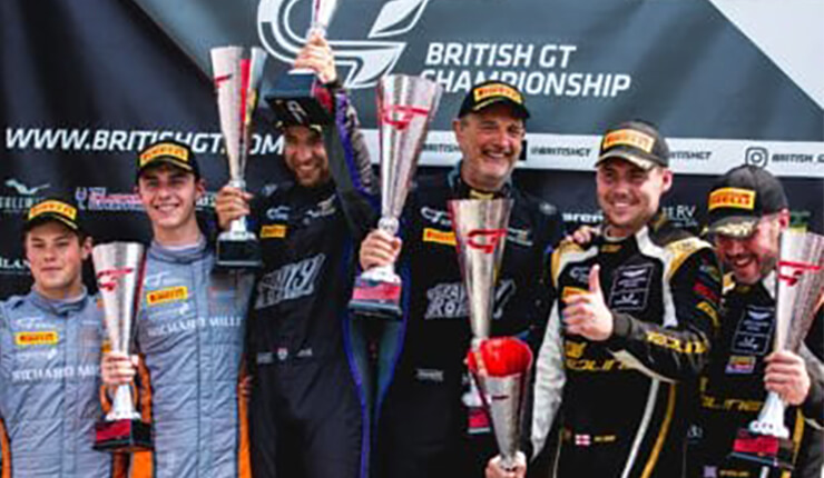 Six men in racing suits and caps holding trophies, celebrating a British GT Championship win.