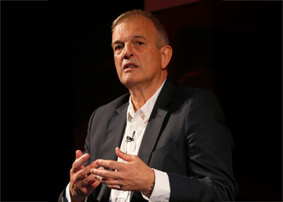 Man in a dark blazer speaking with hands gesturing against a black background.