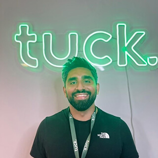 Smiling man with dark hair and beard wearing a black North Face shirt and tuck lanyard, standing in front of a green neon sign that says 'tuck.'