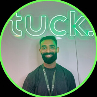 Smiling man with a beard wearing a black shirt and lanyard standing in front of a neon sign that reads 'tuck.'