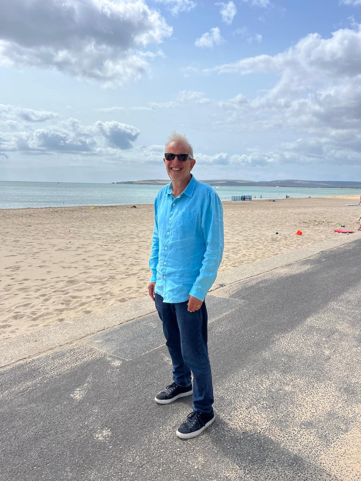 Smiling man wearing sunglasses, a bright blue shirt, and jeans standing on a paved beach walkway with sand and sea in the background under a partly cloudy sky.