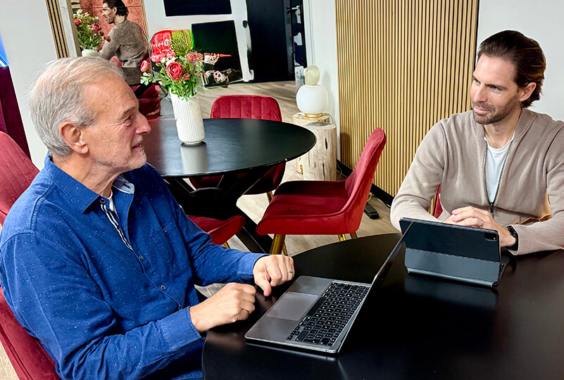 Two men sitting at a black round table in a modern room, one using a laptop and the other using a tablet.