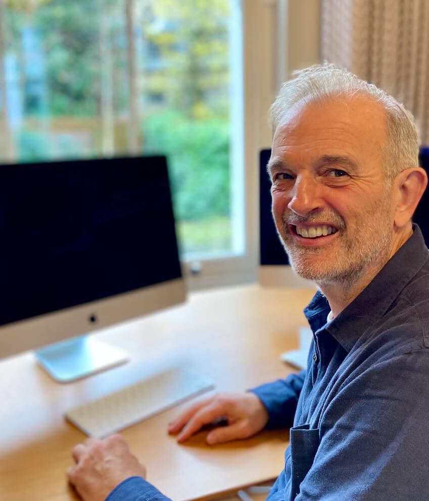 Smiling middle-aged man with gray hair sitting at a desk in front of two computer monitors in a bright room with a window and greenery outside.