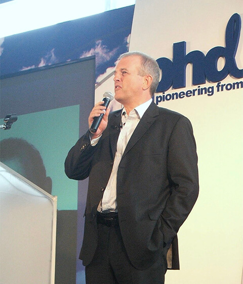 Man in a black suit holding a microphone and speaking at a podium during a presentation.