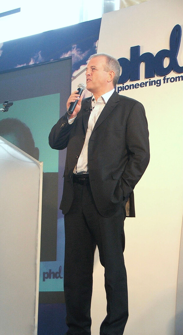 Man in a black suit holding a microphone and speaking at a podium with a background displaying the letters 'phd' and the phrase 'pioneering from'.