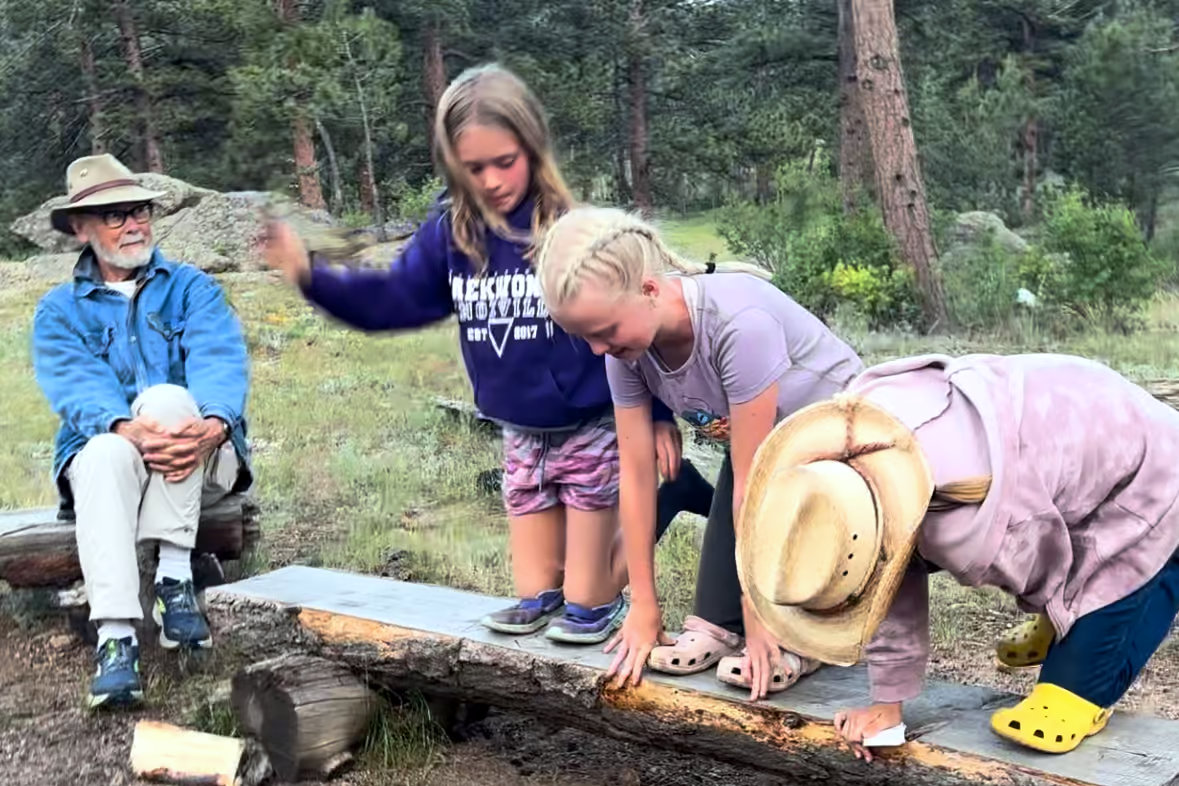 Three children balance playfully on a log bench outdoors while an older man sits nearby watching with a smile.