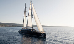 Sleek black sailing yacht with twin masts and full white sails cruising across calm blue water near a distant shoreline under soft daylight.