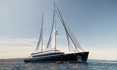 Luxury black sailing yacht with two tall masts and white sails gliding across open water beneath a wide, clear blue sky.