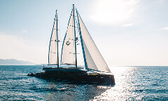 Luxury black sailing yacht with twin masts and white sails marked by a star emblem, cruising on sparkling sunlit water.