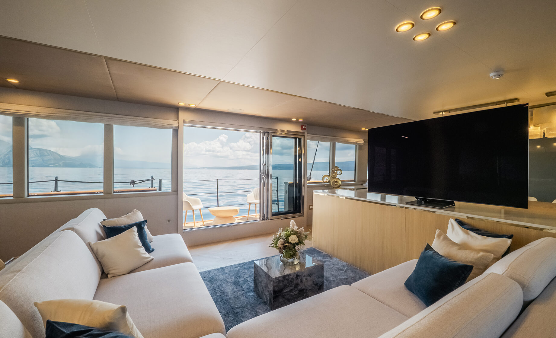 Modern yacht lounge with white sofas and blue accent pillows, a large flat-screen TV, and panoramic windows overlooking the sea and distant mountains.