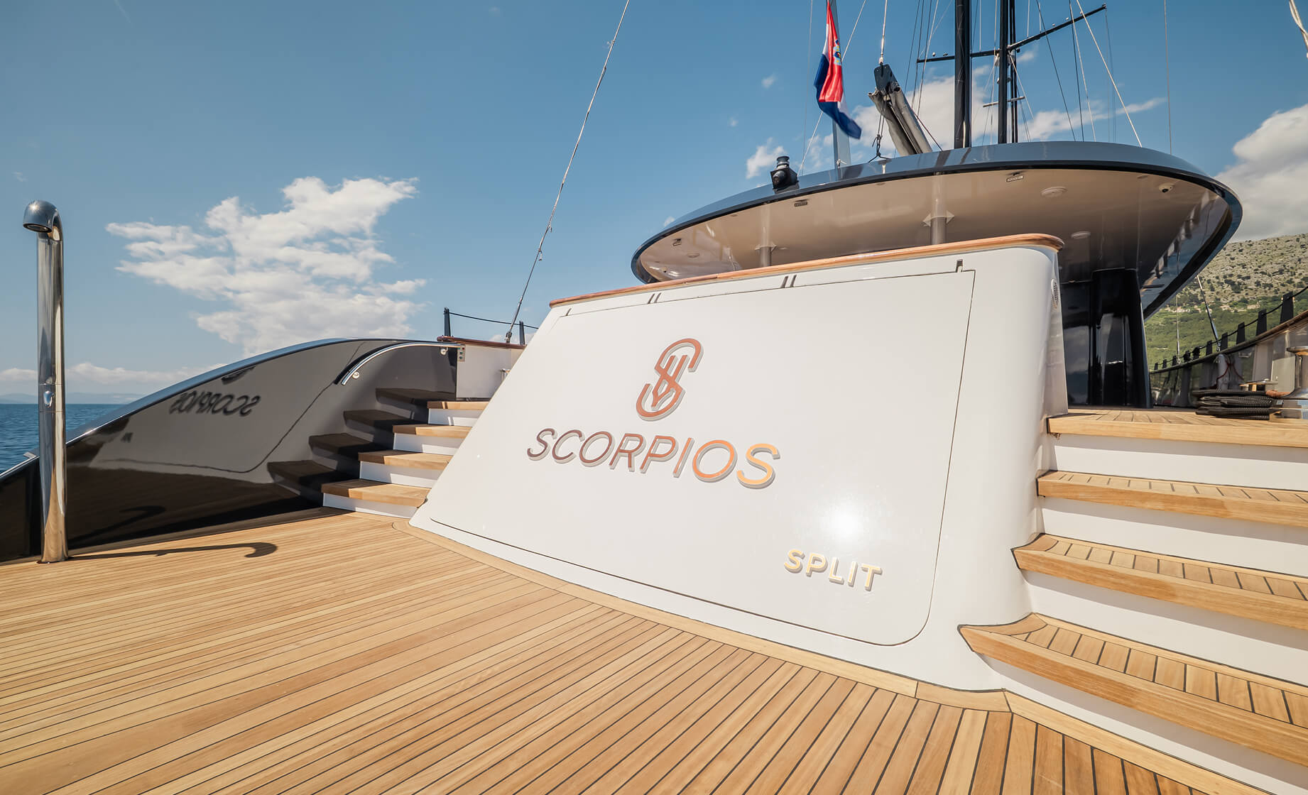 Teak deck of the luxury yacht “Scorpios” with wooden stairs on both sides, a curved white structure displaying the Scorpios logo and “Split,” under a clear blue sky.