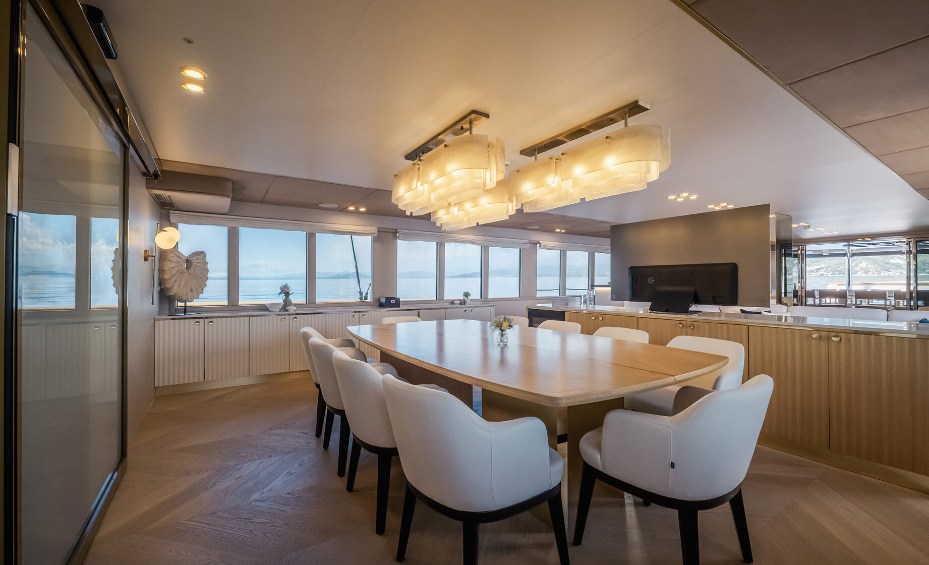 Elegant yacht dining area with long table, upholstered chairs, warm wood finishes, and panoramic sea views through large windows.