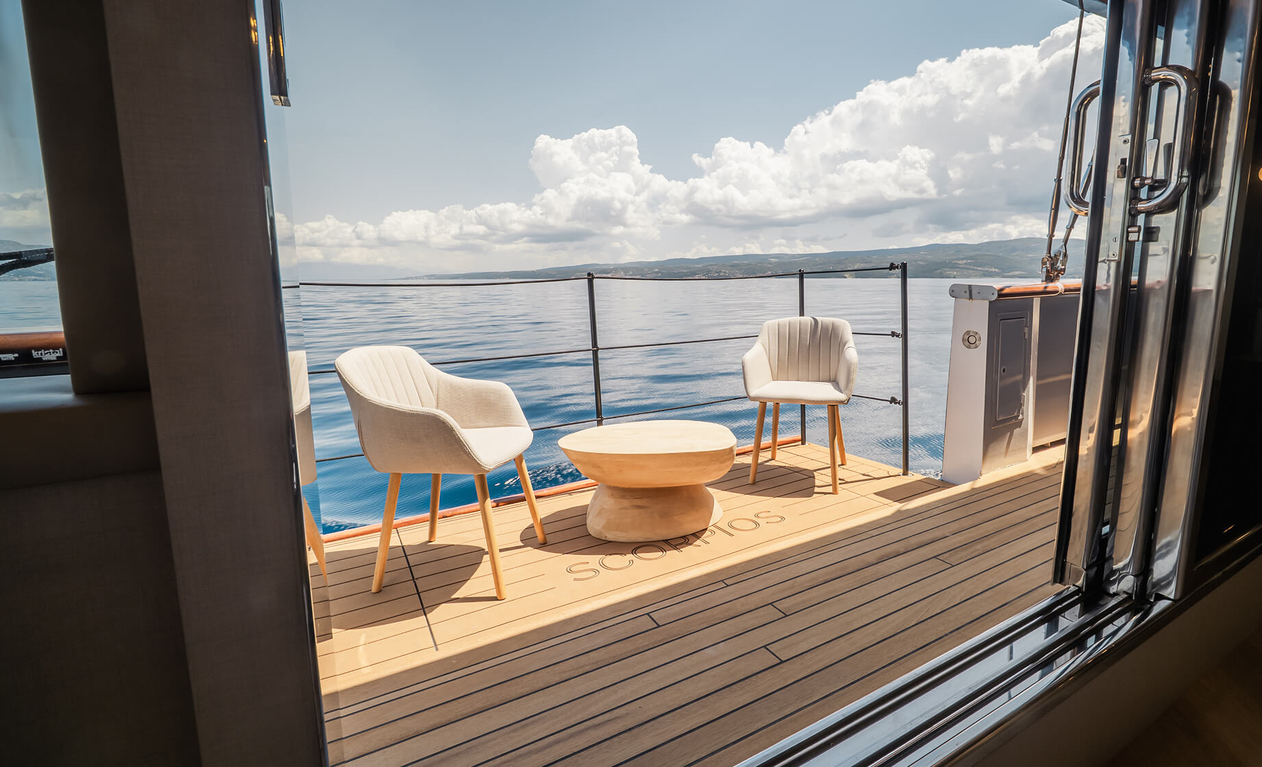 View from inside a luxury yacht cabin onto a private deck with two upholstered chairs and a round wooden table, overlooking calm sea and distant mountains under a bright sky.