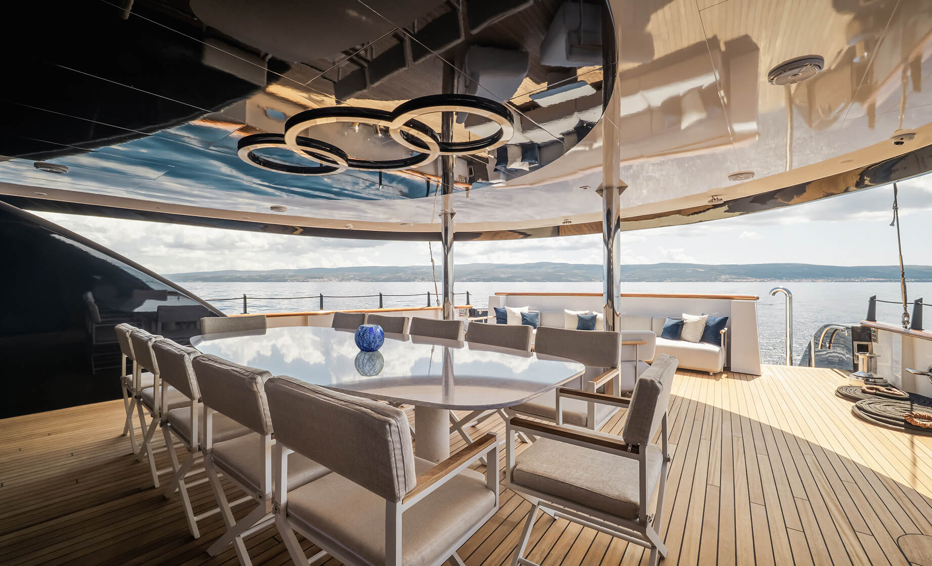 Luxury yacht dining area with a large oval table and cushioned chairs on a teak deck, mirrored ceiling with circular light fixtures, and panoramic sea views in the background.