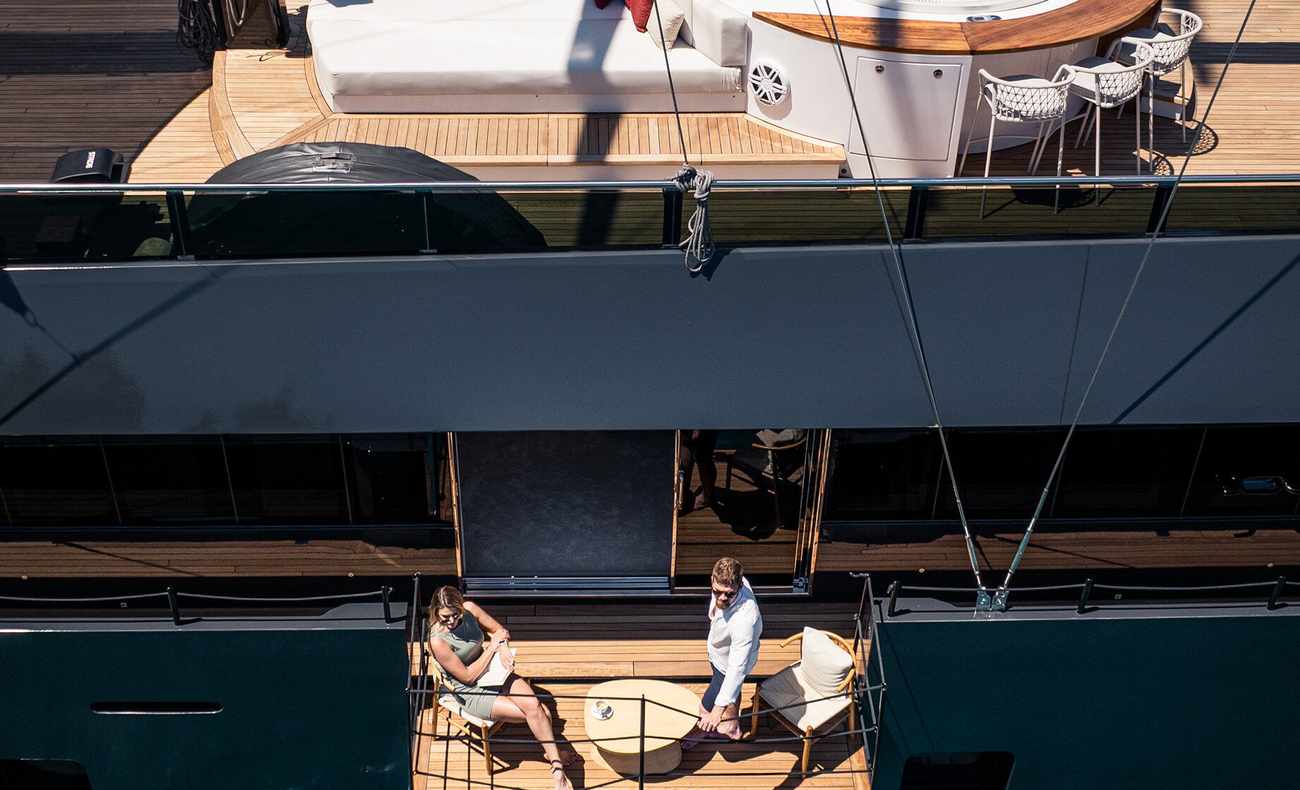 Top-down view of a couple relaxing on a private balcony deck of a luxury yacht, seated around a small table above turquoise water.