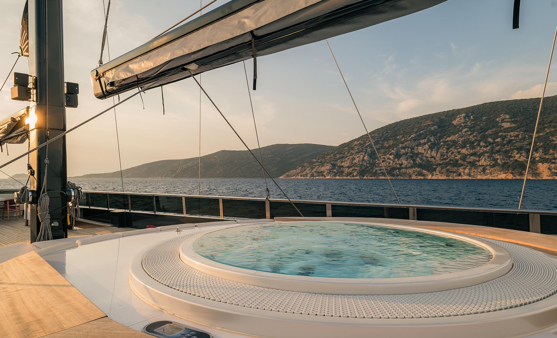 Outdoor yacht deck with a built-in jacuzzi, overlooking calm sea and rocky hills at sunset.