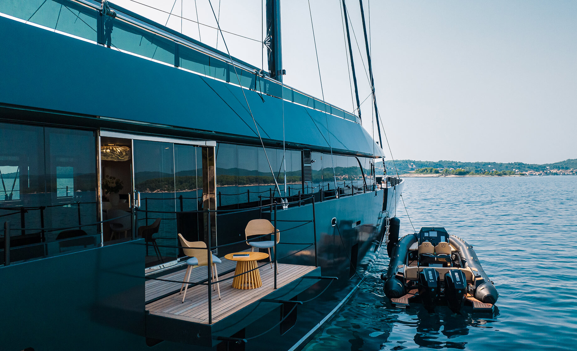 Side view of a modern luxury sailing yacht with a private deck terrace, outdoor chairs and table, and an inflatable tender boat moored alongside on calm water near the coast.