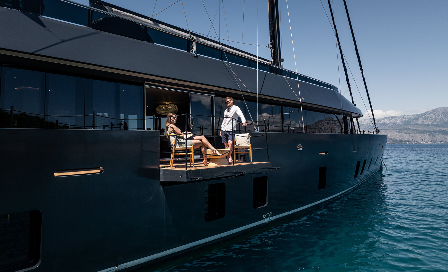 Couple relaxing on a private terrace of a luxury sailing yacht, seated on deck chairs above calm blue water with mountains in the background.