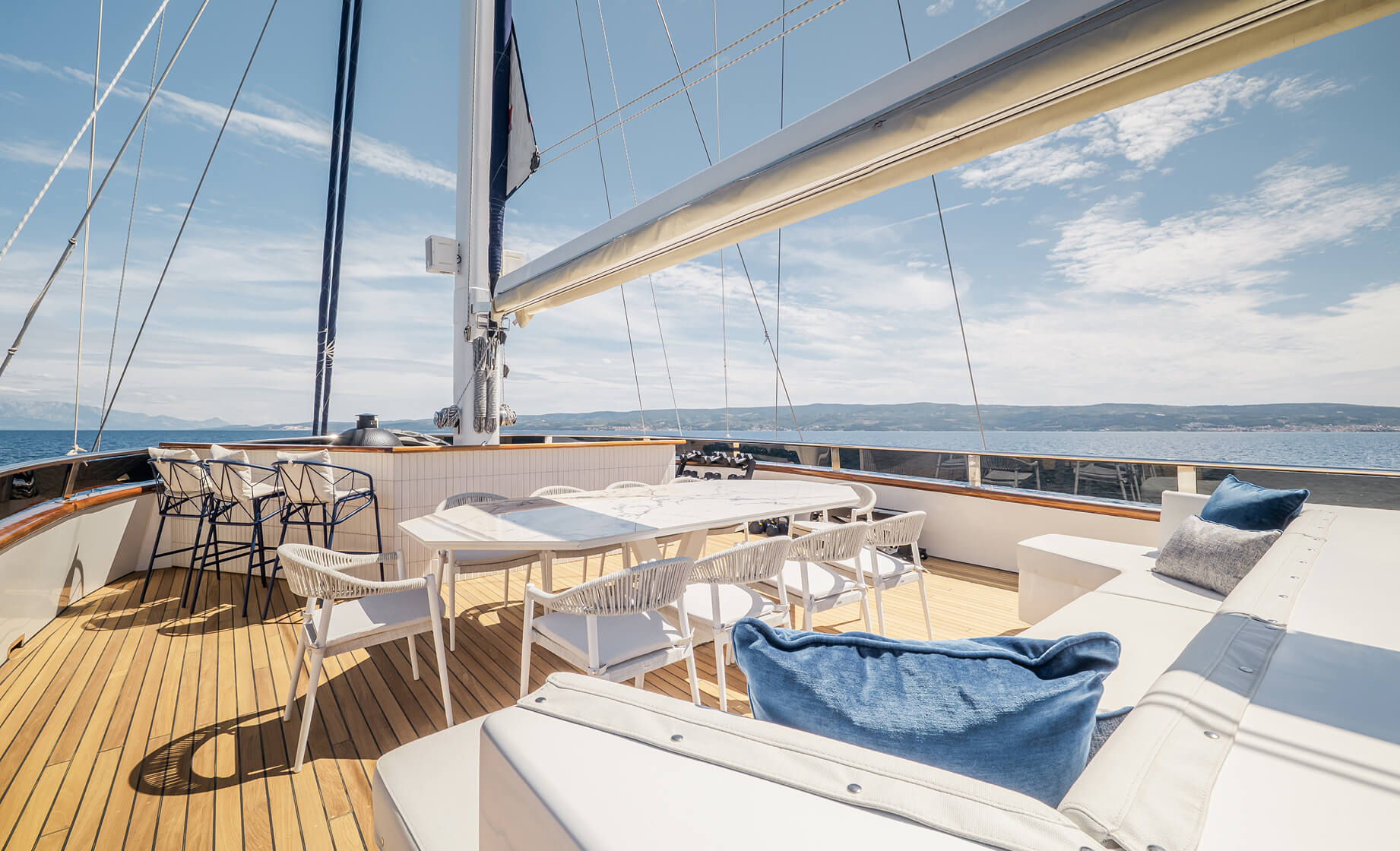 Open-air yacht deck with a dining table and white chairs, bar stools, and cushioned seating, set against a backdrop of calm sea and distant coastline under a clear blue sky.