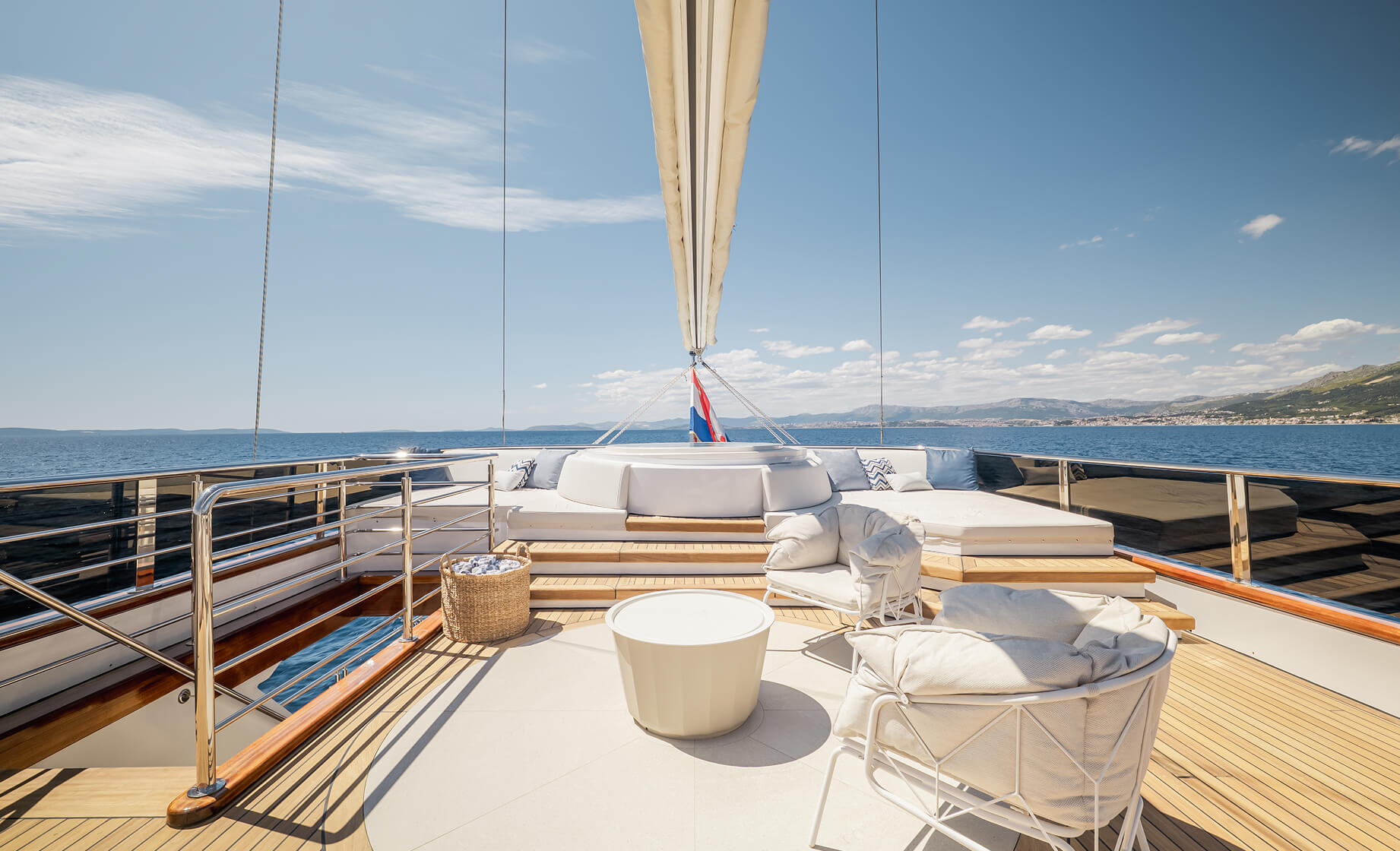 Bright yacht sundeck with round cushioned seating, lounge area and small table, overlooking calm blue sea and distant coastline beneath a clear sky.