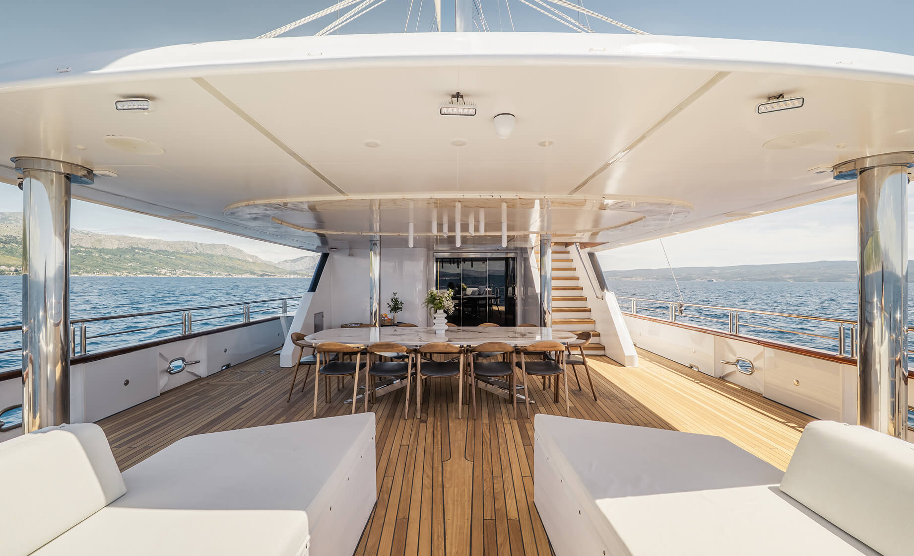 Spacious covered yacht deck with a long dining table and wooden chairs, teak flooring, stainless steel pillars, and open sea views on both sides under clear daylight.