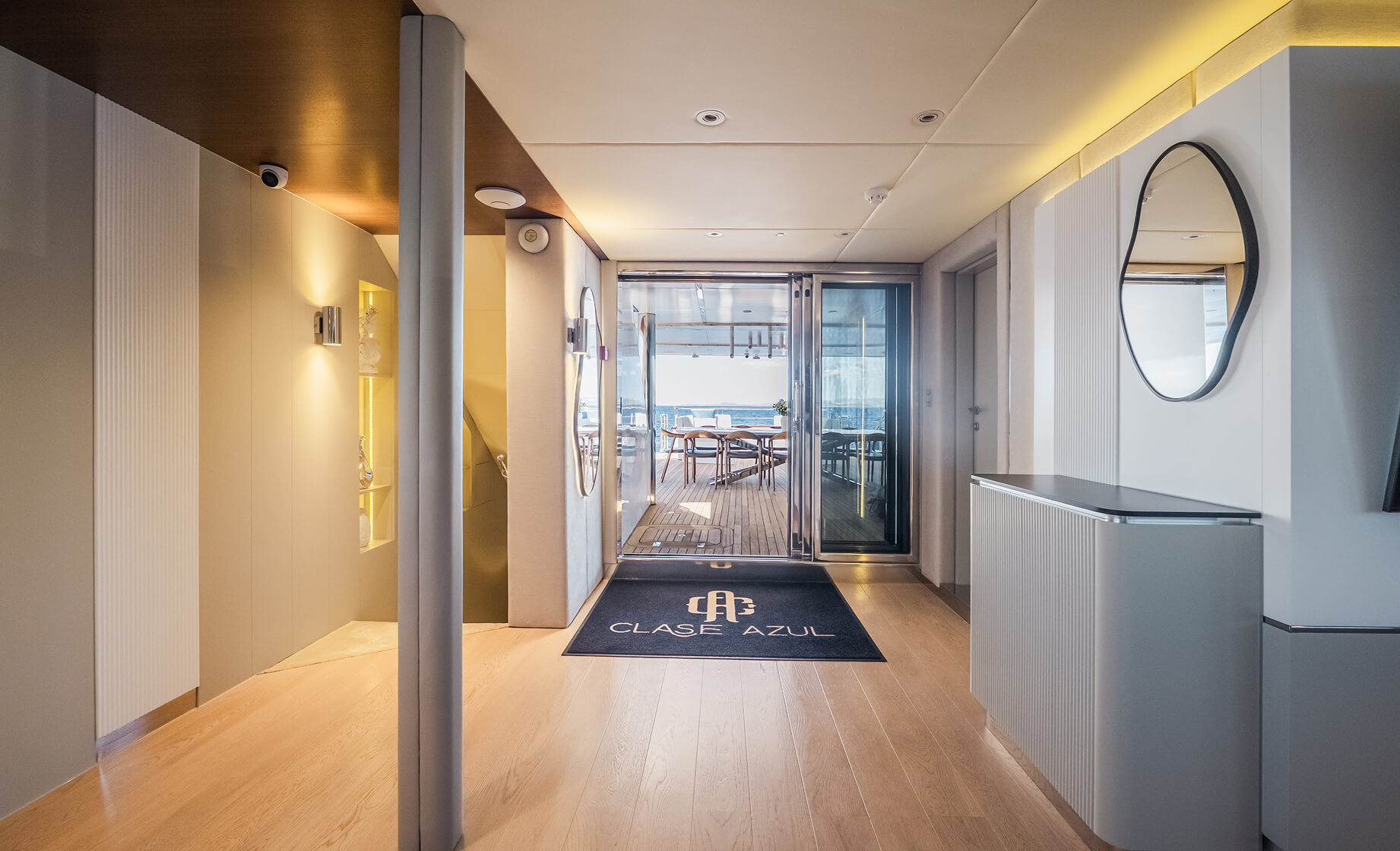 Modern yacht interior hallway with light wood flooring, minimalist décor, a “Clase Azul” floor mat, and glass doors leading to an outdoor deck dining area with sea views.