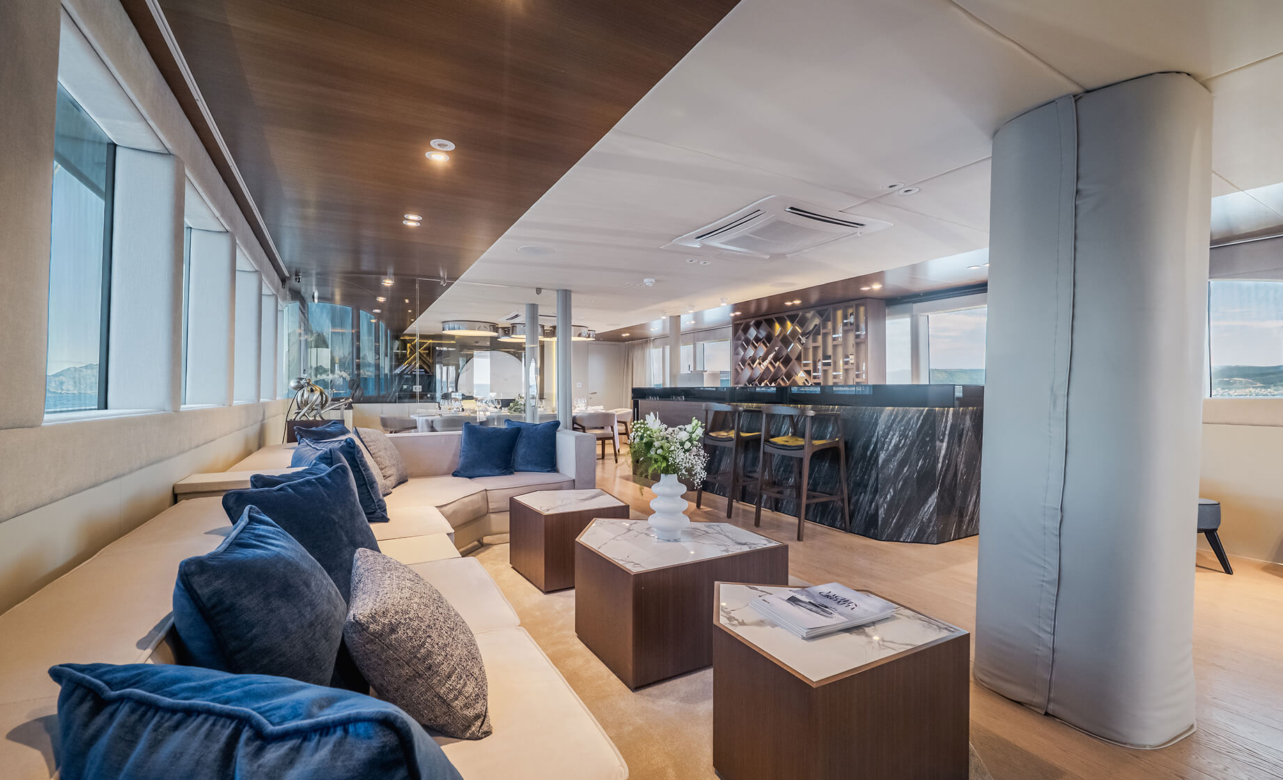 Spacious luxury yacht lounge with cream sectional sofa and blue accent pillows, marble-top coffee tables, and a modern bar area with stools and wine display in the background.