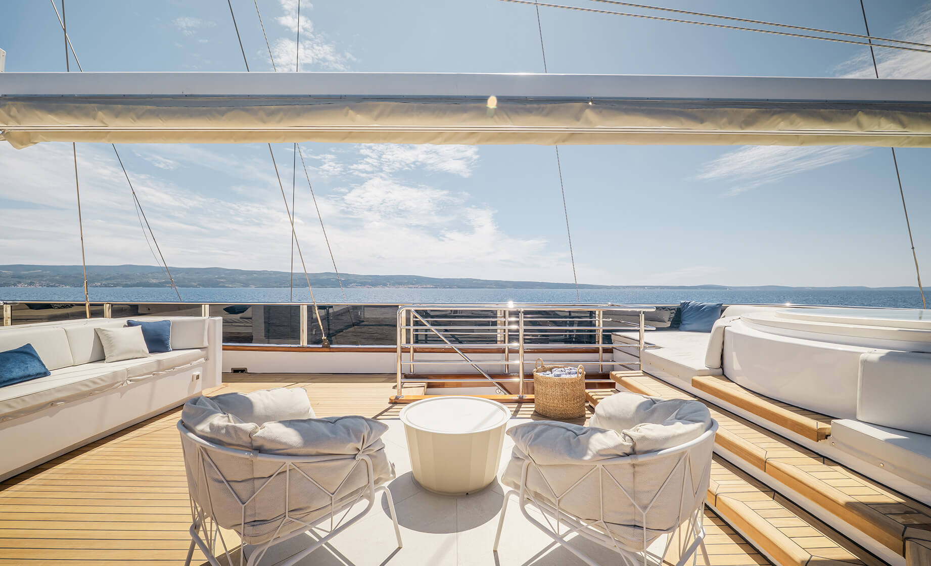 Sunny yacht sundeck with cushioned lounge seating, two round chairs and a small table, a jacuzzi, and open sea views beneath a clear blue sky.
