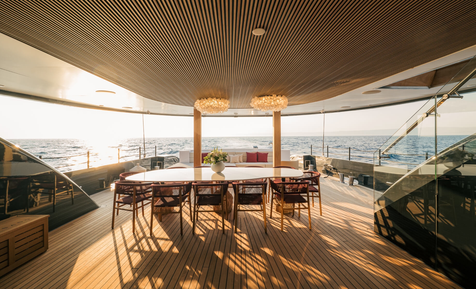 Spacious yacht deck dining area at sunset with a large table and chairs, wooden flooring, modern lighting, and panoramic ocean views.