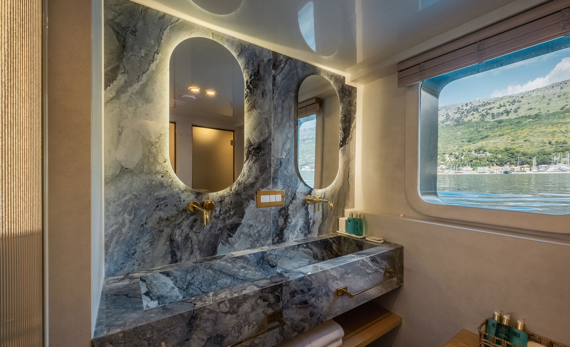 Luxury yacht bathroom with a marble vanity and backsplash, dual oval mirrors with backlighting, gold fixtures, and a window overlooking the sea and green hills.
