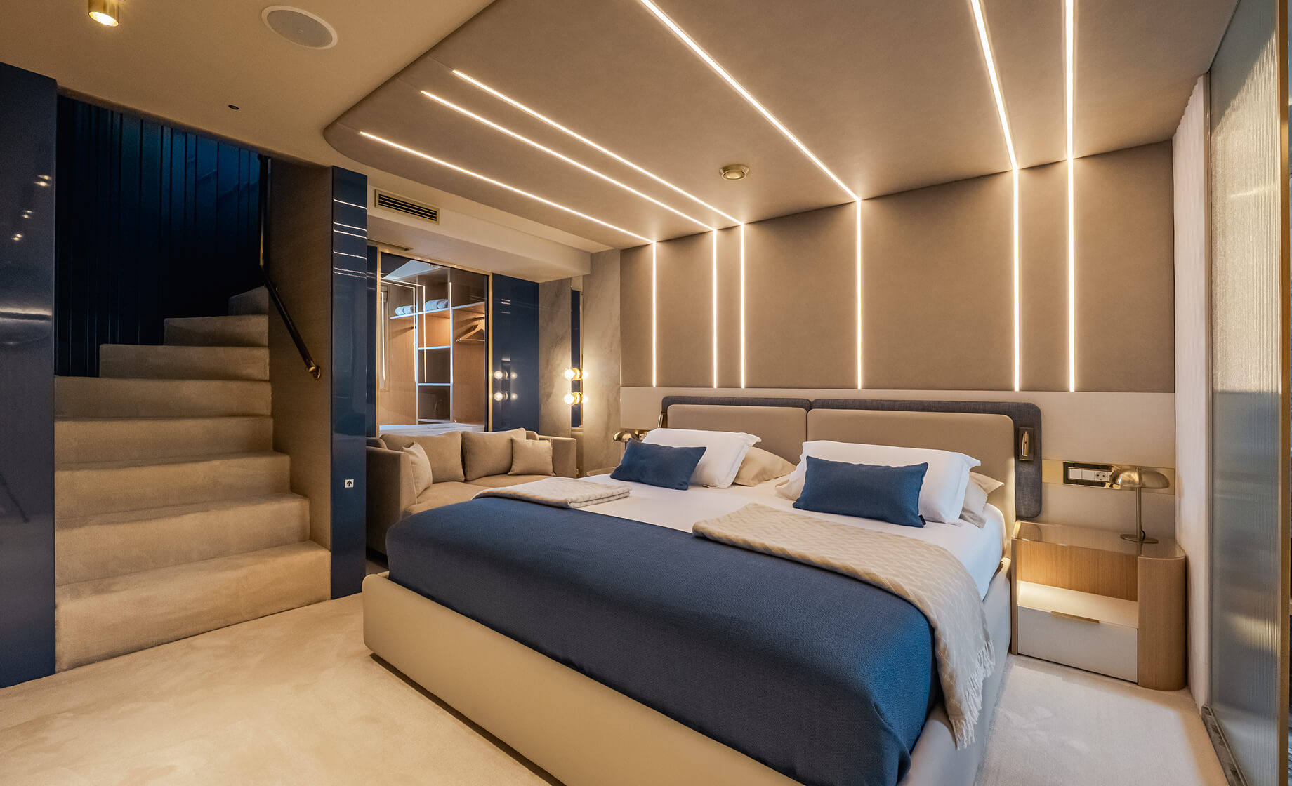View from inside a luxury yacht cabin onto a private deck with two upholstered chairs and a round wooden table, overlooking calm sea and distant mountains under a bright sky.