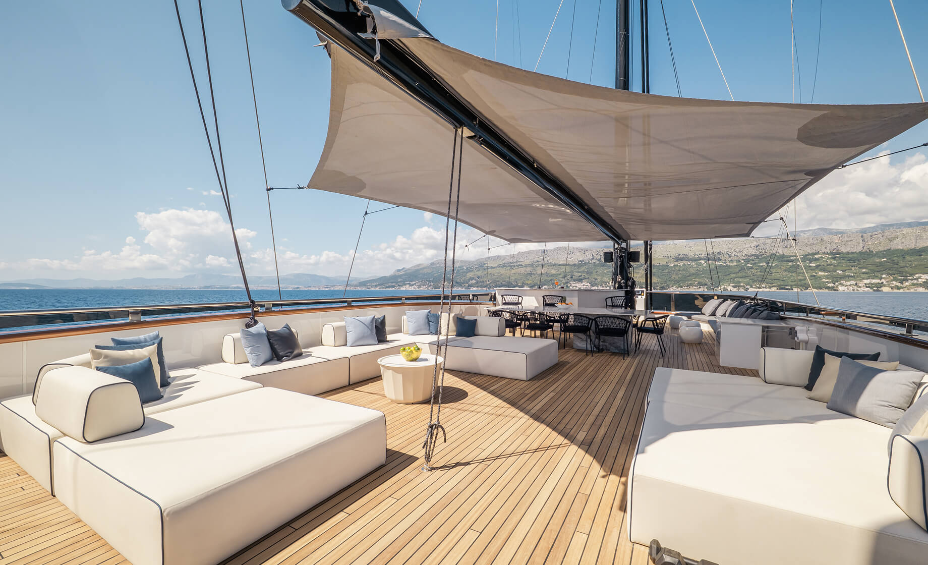 View from inside a luxury yacht cabin onto a private deck with two upholstered chairs and a round wooden table, overlooking calm sea and distant mountains under a bright sky.
