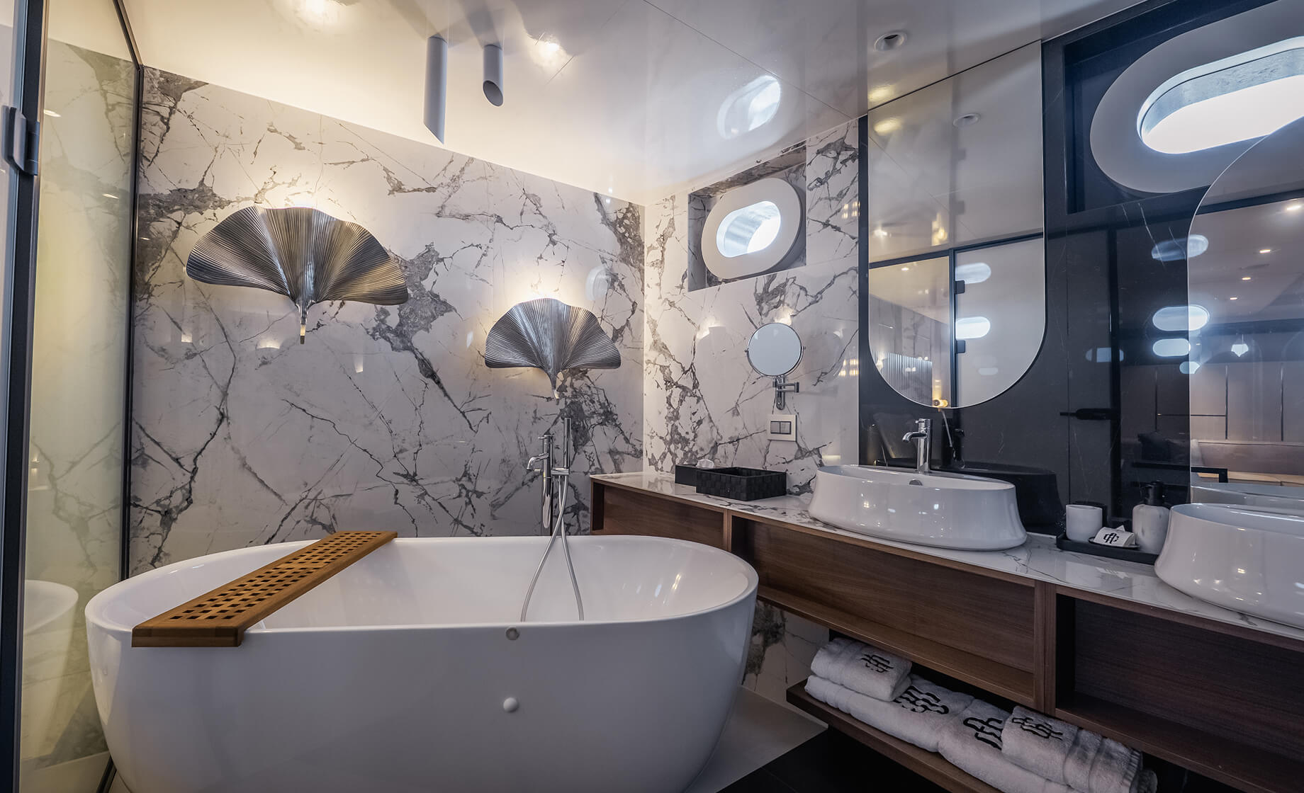 Elegant yacht bathroom with a freestanding bathtub, marble walls, wooden vanity with vessel sinks, oval mirrors, and soft ambient lighting.