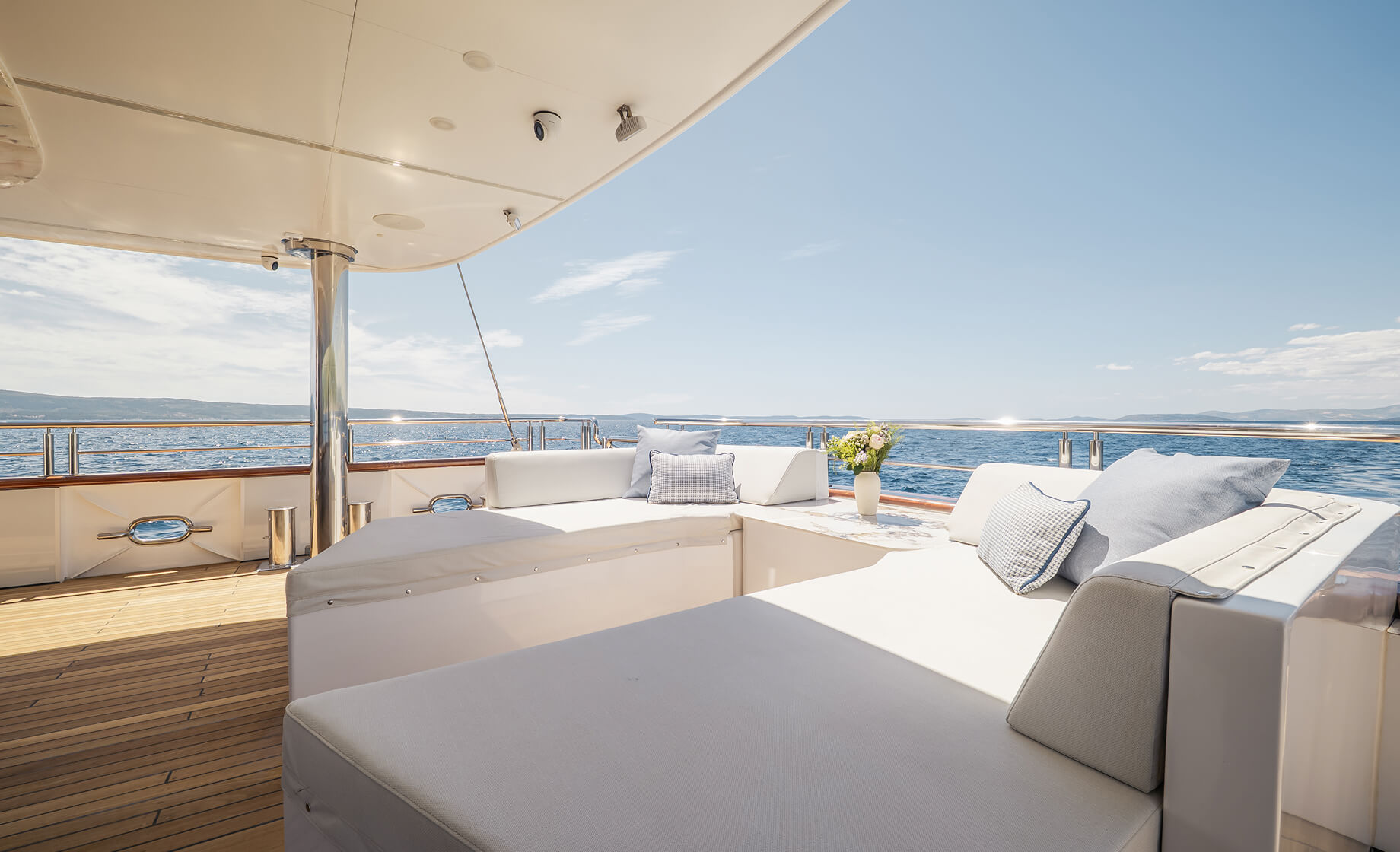 Luxury yacht outdoor lounge with white cushioned seating, decorative pillows and a small table with flowers, overlooking calm blue sea under a clear sky.