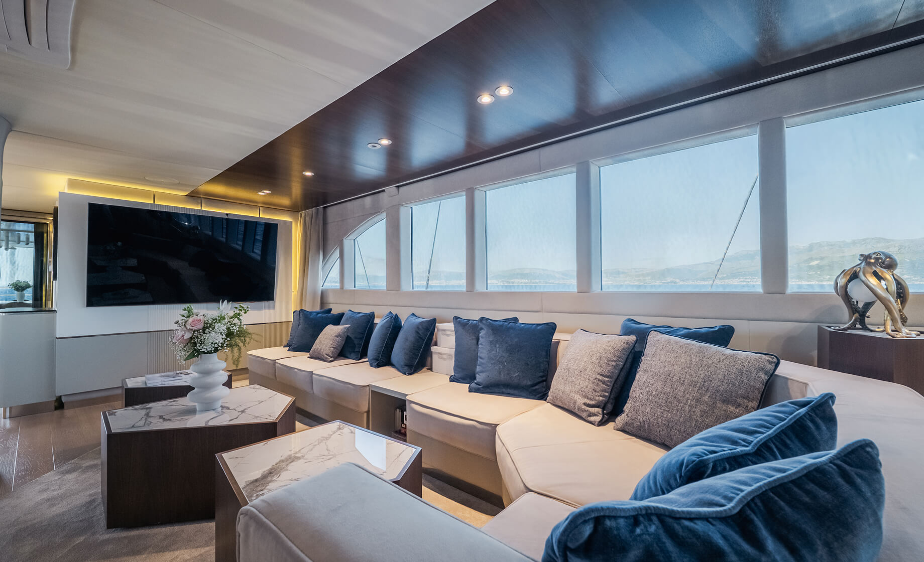 Modern yacht lounge with long cream sofa and blue accent pillows, marble-top coffee tables, large flat-screen TV, and panoramic windows overlooking the sea.