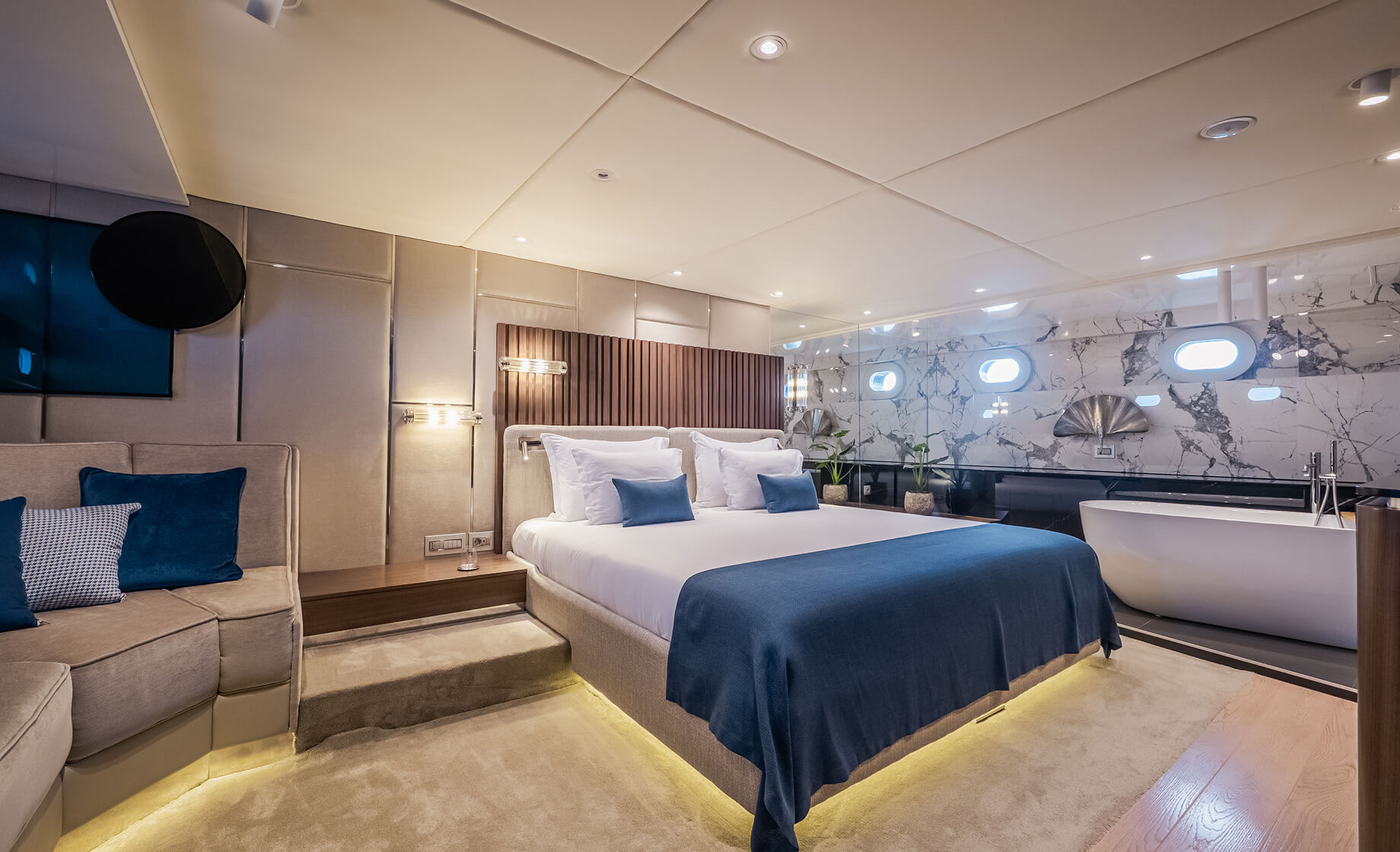 Luxurious yacht master cabin with a king-size bed, blue accents, soft ambient lighting, marble wall finishes, and a freestanding bathtub beside oval windows.