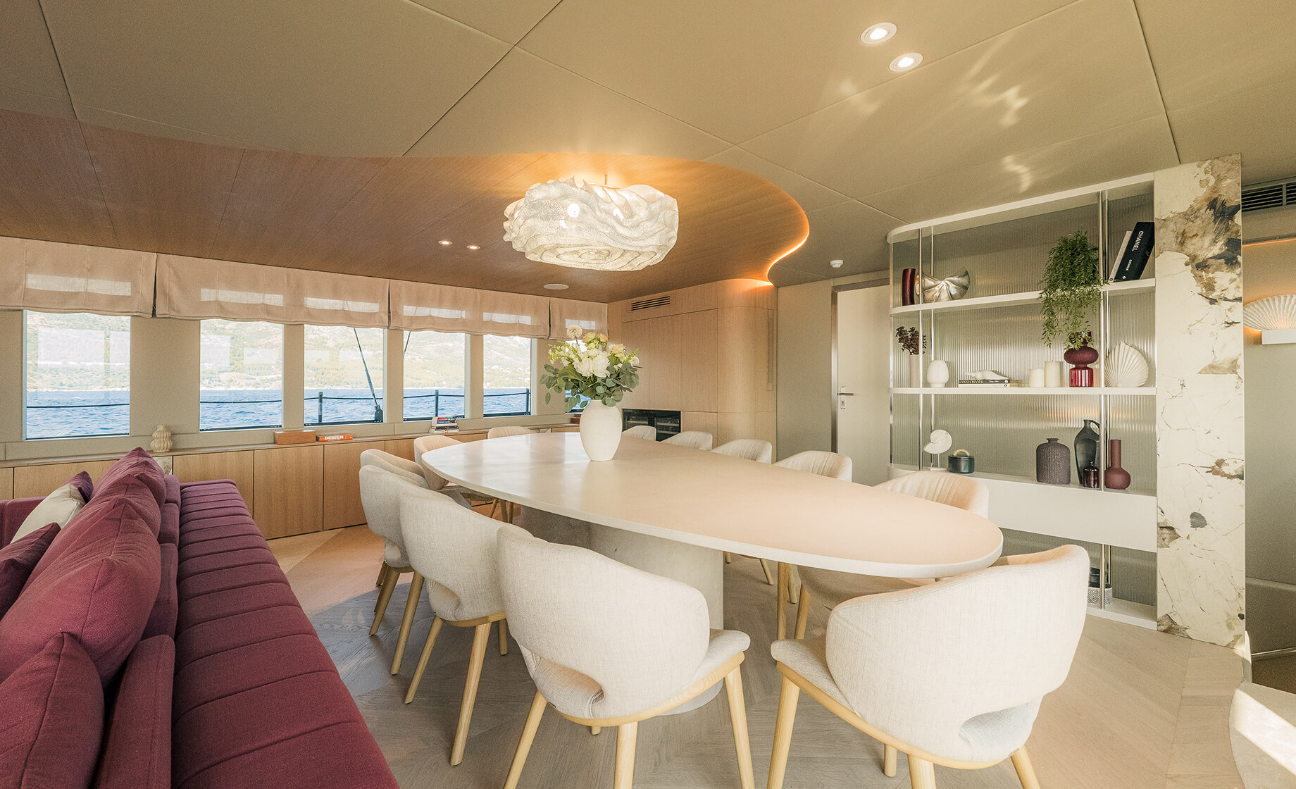 Modern yacht dining and lounge area with an oval table, upholstered chairs, built-in shelving, and soft ambient lighting overlooking the sea.