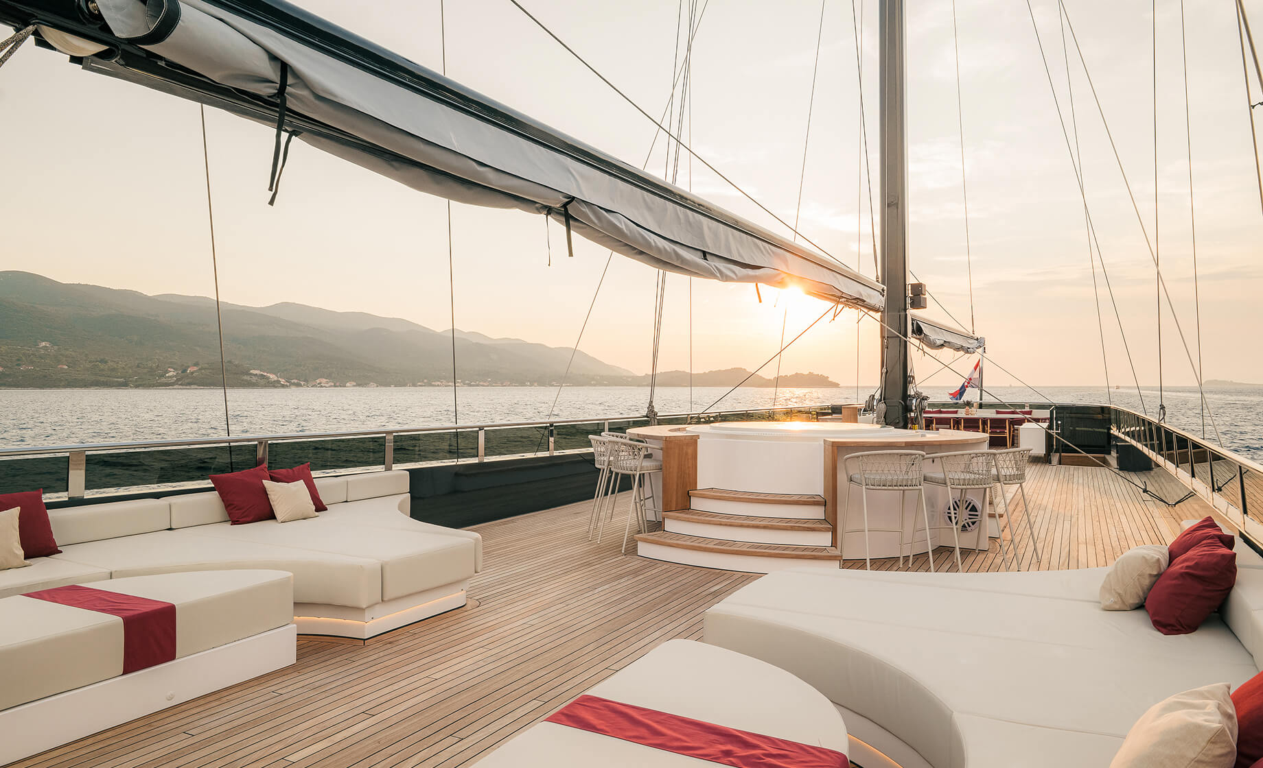 Luxury yacht sun deck at sunset with spacious lounge seating, a central bar area, and panoramic views of the sea and mountains.