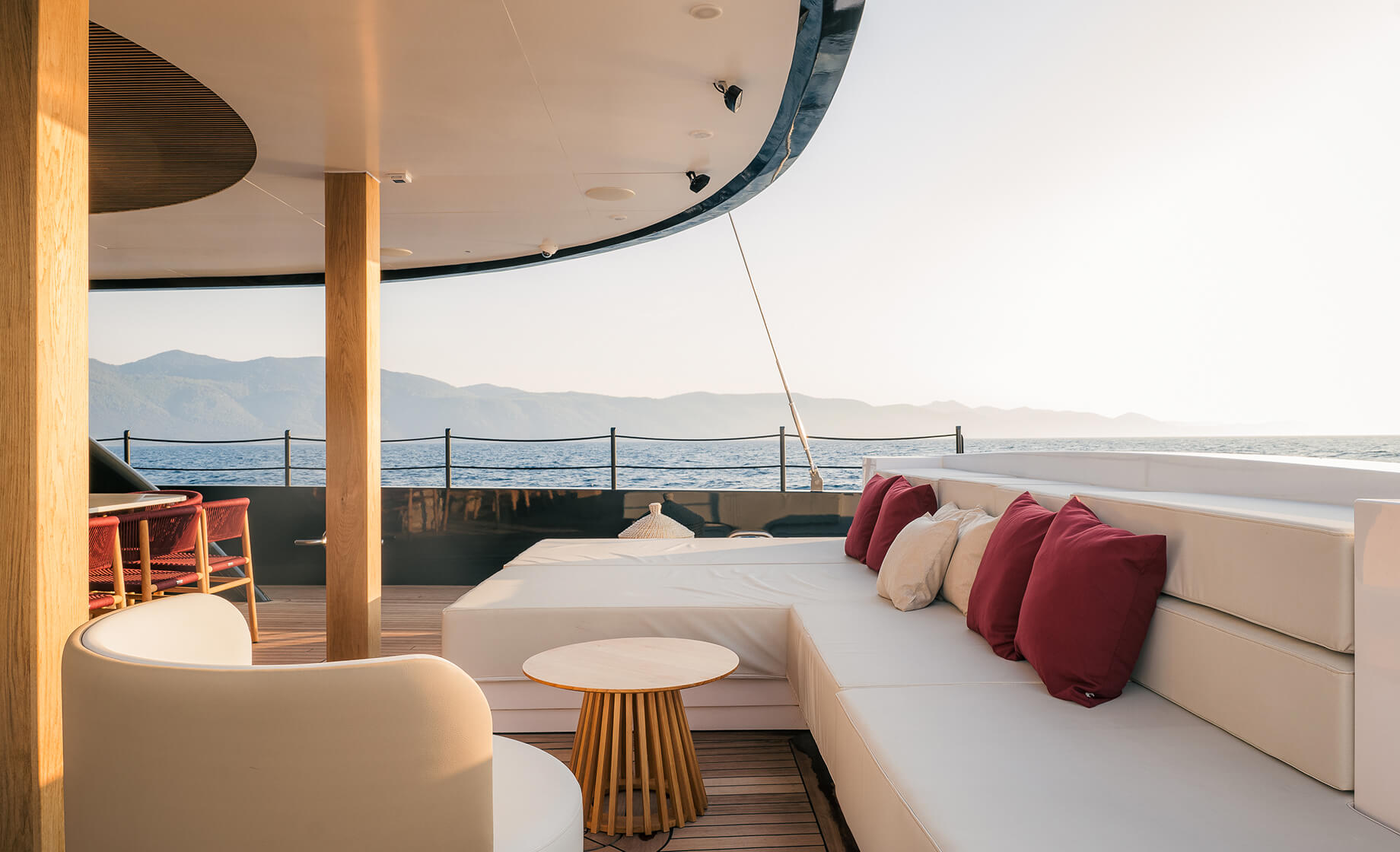 Luxury yacht lounge deck with curved white seating, red accent pillows, and panoramic sea and mountain views at sunset.