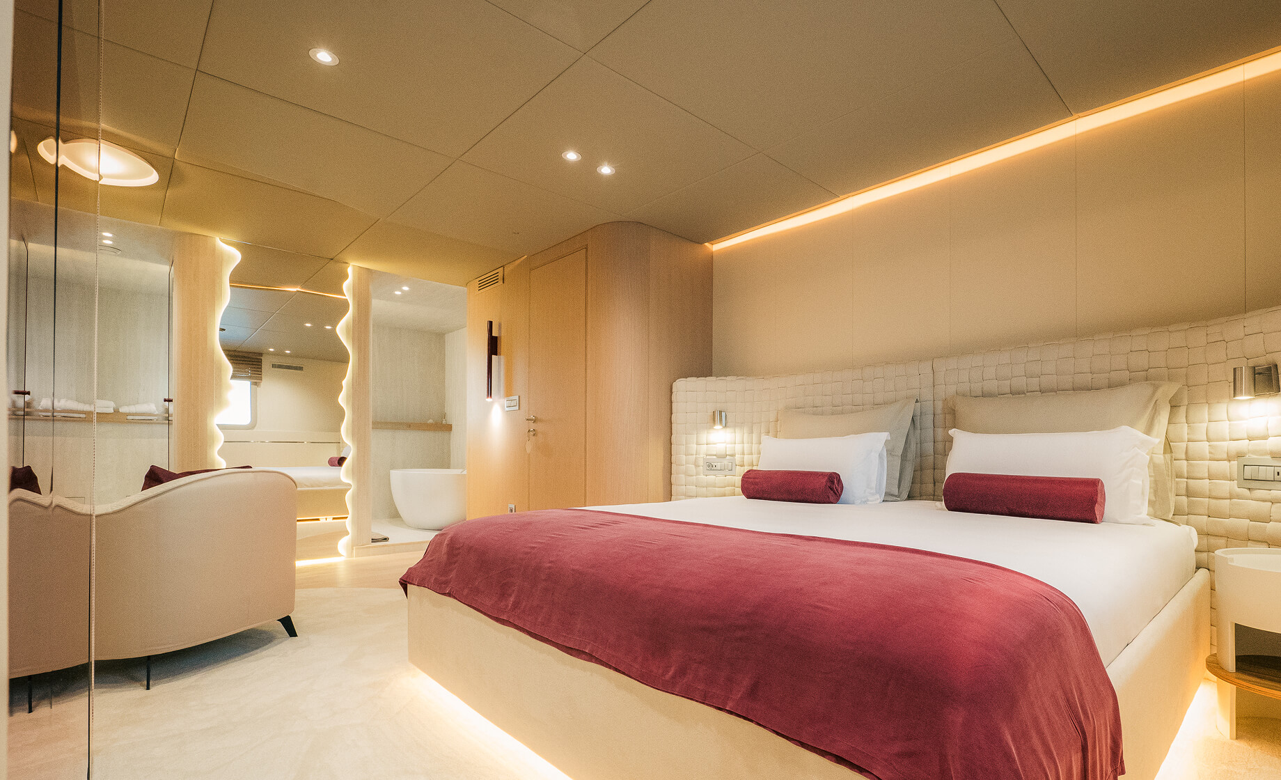 Luxury yacht master bedroom with a large upholstered bed, soft ambient lighting, red accent throw, and a modern en-suite bathroom visible through glass panels.