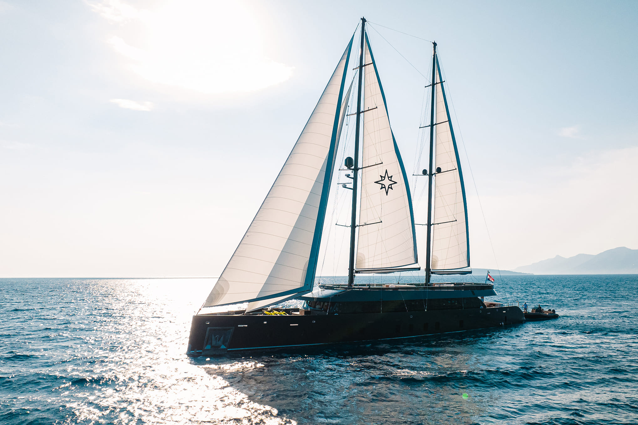 Luxury twin-masted sailing yacht with white sails cruising on sunlit blue sea, coastline and distant mountains visible on the horizon.