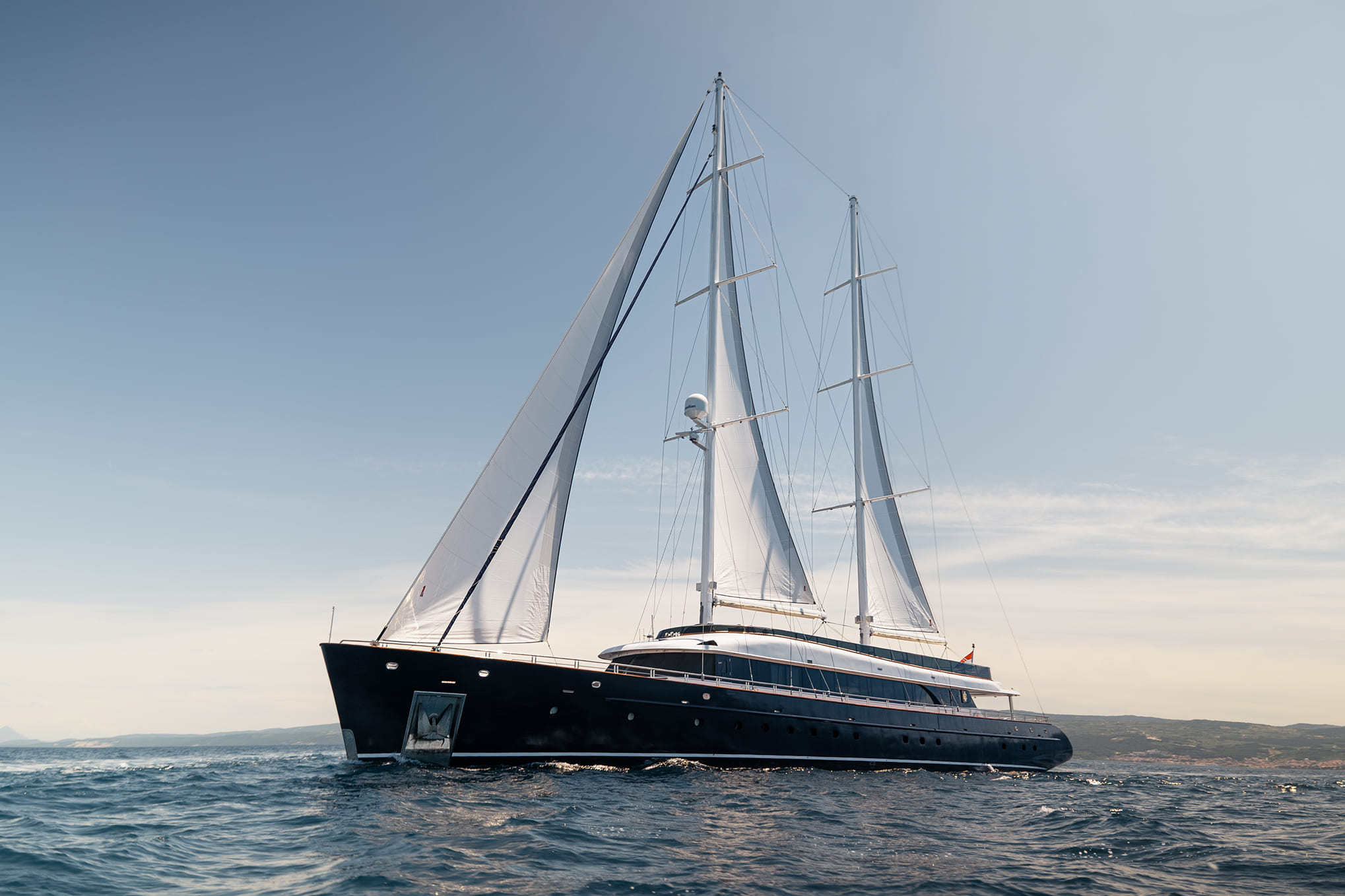 Luxury black superyacht with twin masts and full white sails gliding across open sea under a clear blue sky, with distant coastline on the horizon.