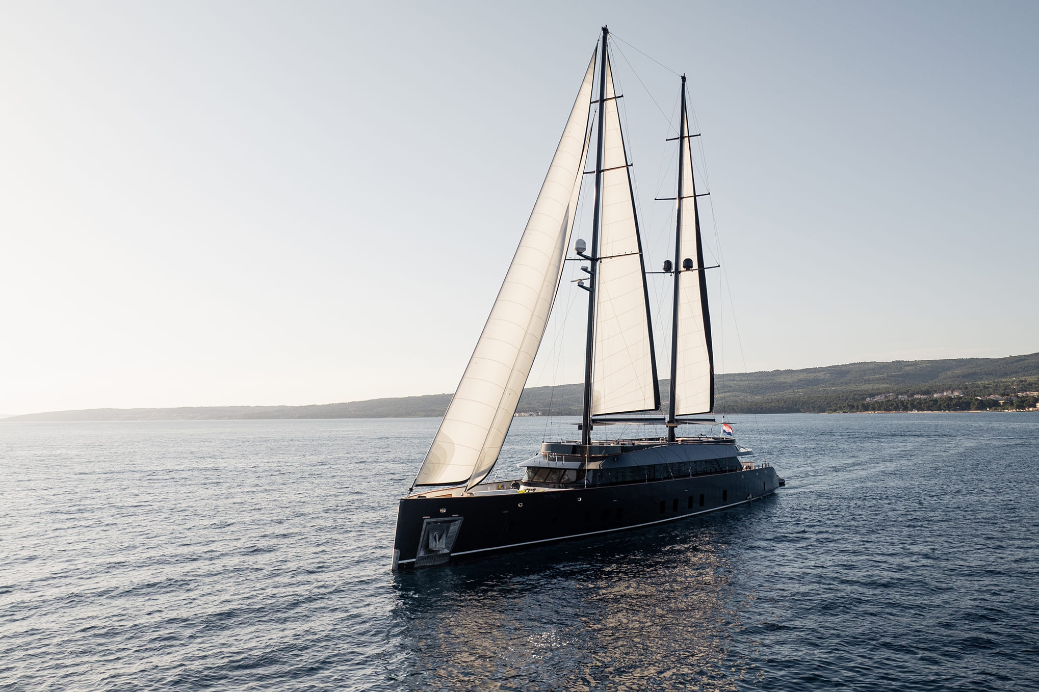 Luxury black sailing yacht with two tall masts and white sails cruising on calm blue sea near a green coastline at sunset.