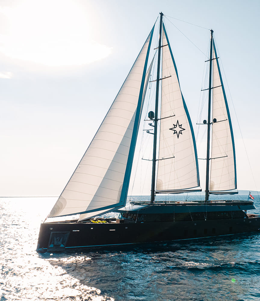 Luxury black sailing yacht with twin masts and white sails marked by a star emblem, cruising on sparkling sunlit water.