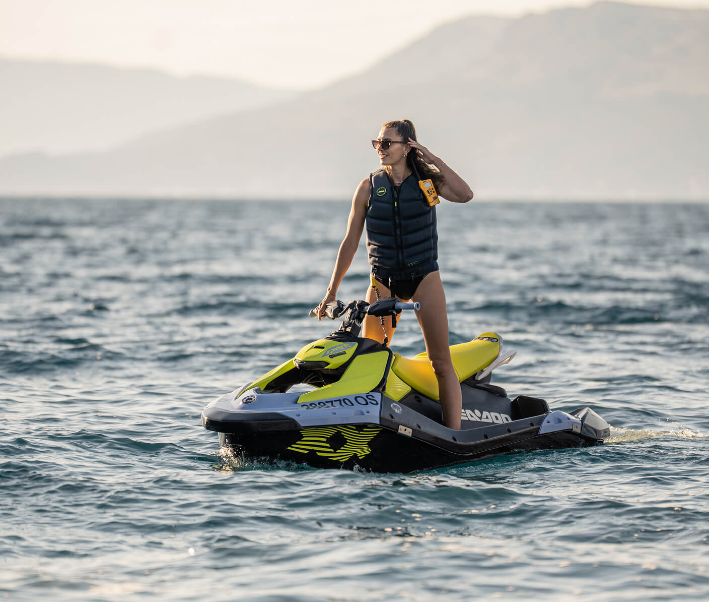 Woman wearing a life vest and sunglasses standing on a yellow jet ski in open water, with mountains in the background at sunset.