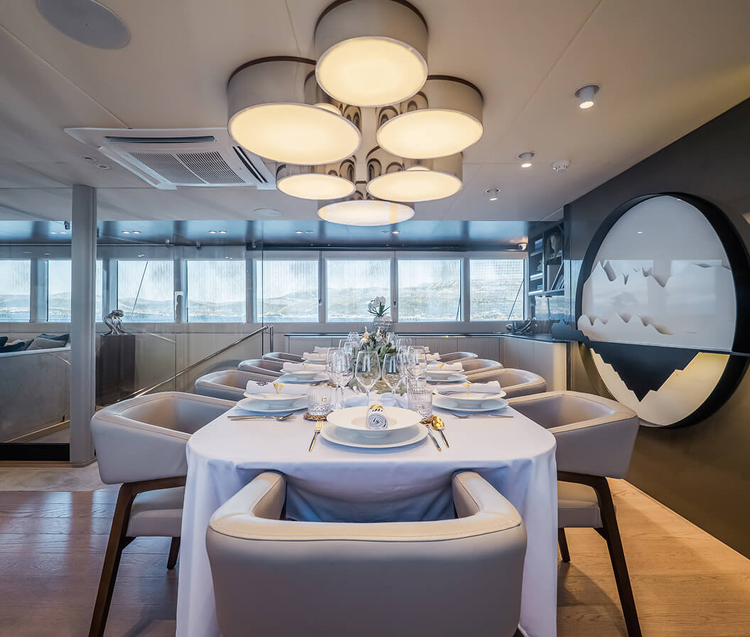 Elegant yacht dining room with a long table set for a formal meal, upholstered chairs, modern circular ceiling lights, and large panoramic windows overlooking the sea.