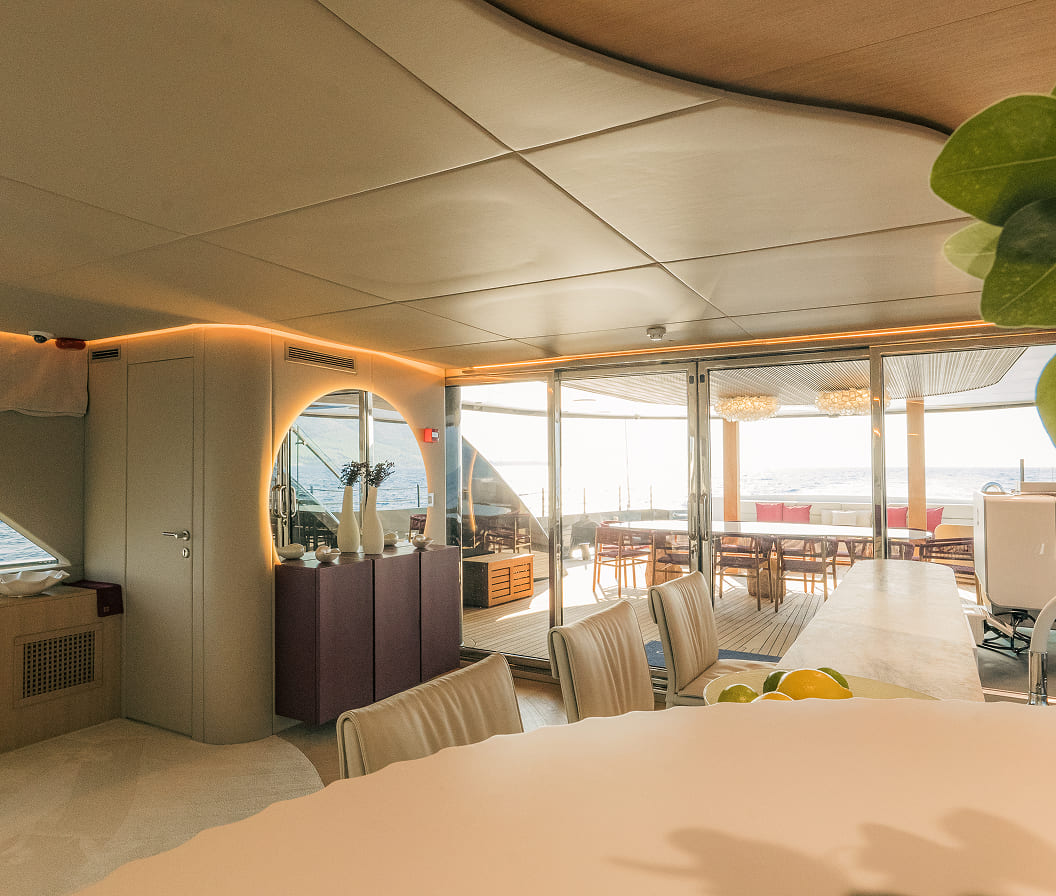 Modern yacht interior dining area with soft ambient lighting, sliding glass doors opening to an outdoor deck, and ocean views in the background.