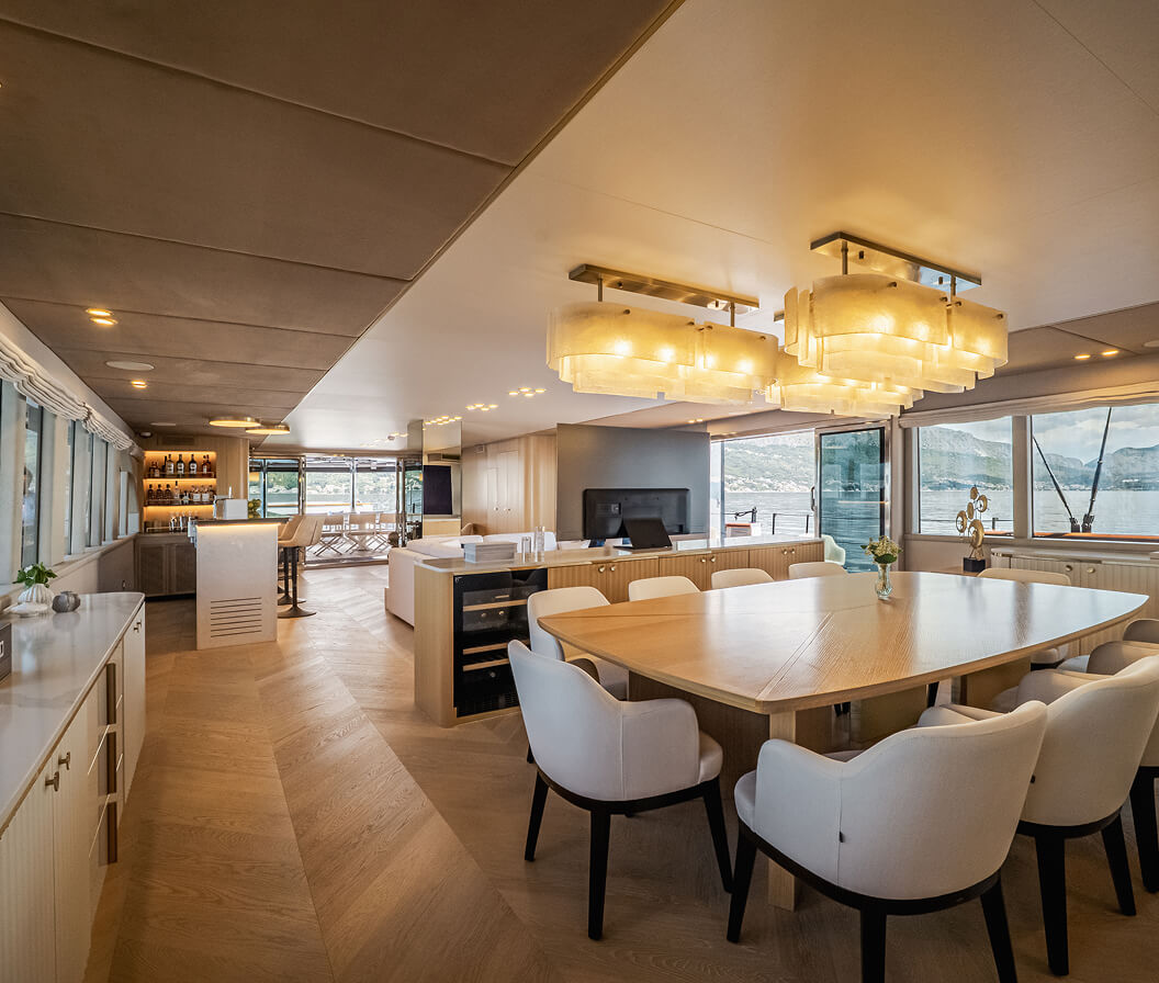 Luxurious yacht interior with spacious dining area, modern chandelier lighting, cream upholstered chairs, and panoramic ocean views through large windows.