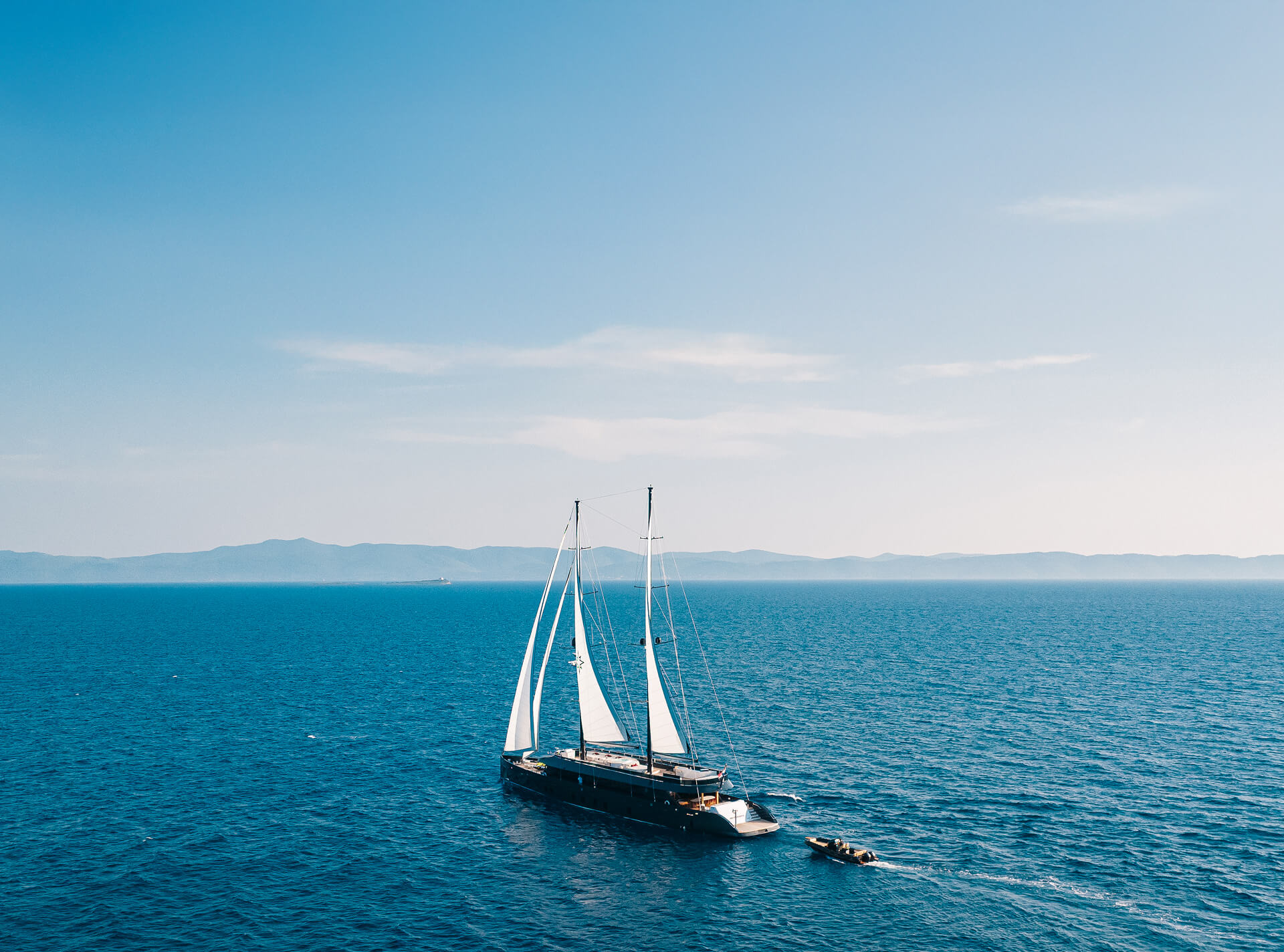 Aerial view of a luxury sailing yacht with white sails cruising across open blue sea, accompanied by a small tender boat.